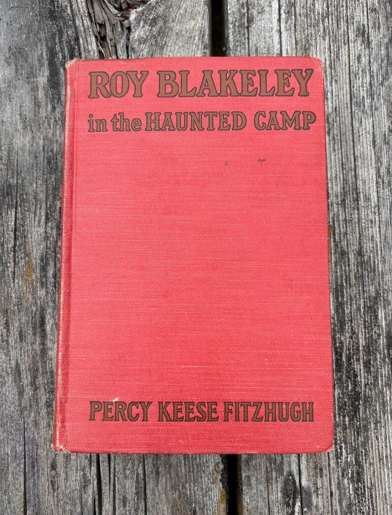 May include: A red hardcover book titled Roy Blakeley in the Haunted Camp by Percy Keese Fitzhugh. The front cover features dark brown text printed on a textured red fabric surface, with the title at the top and the author name at the bottom.