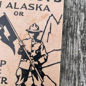 K&ouml;nnte beinhalten: Ein Vintage-Buchcover mit einem hellbraunen, strukturierten Hintergrund und schwarzen Tintenillustrationen. Der Text lautet BOY SCOUT'S IN ALASKA OR. Eine zentrale Grafik zeigt einen Pfadfinder mit breitkrempigem Hut, der einen Fahnenmast h&auml;lt.