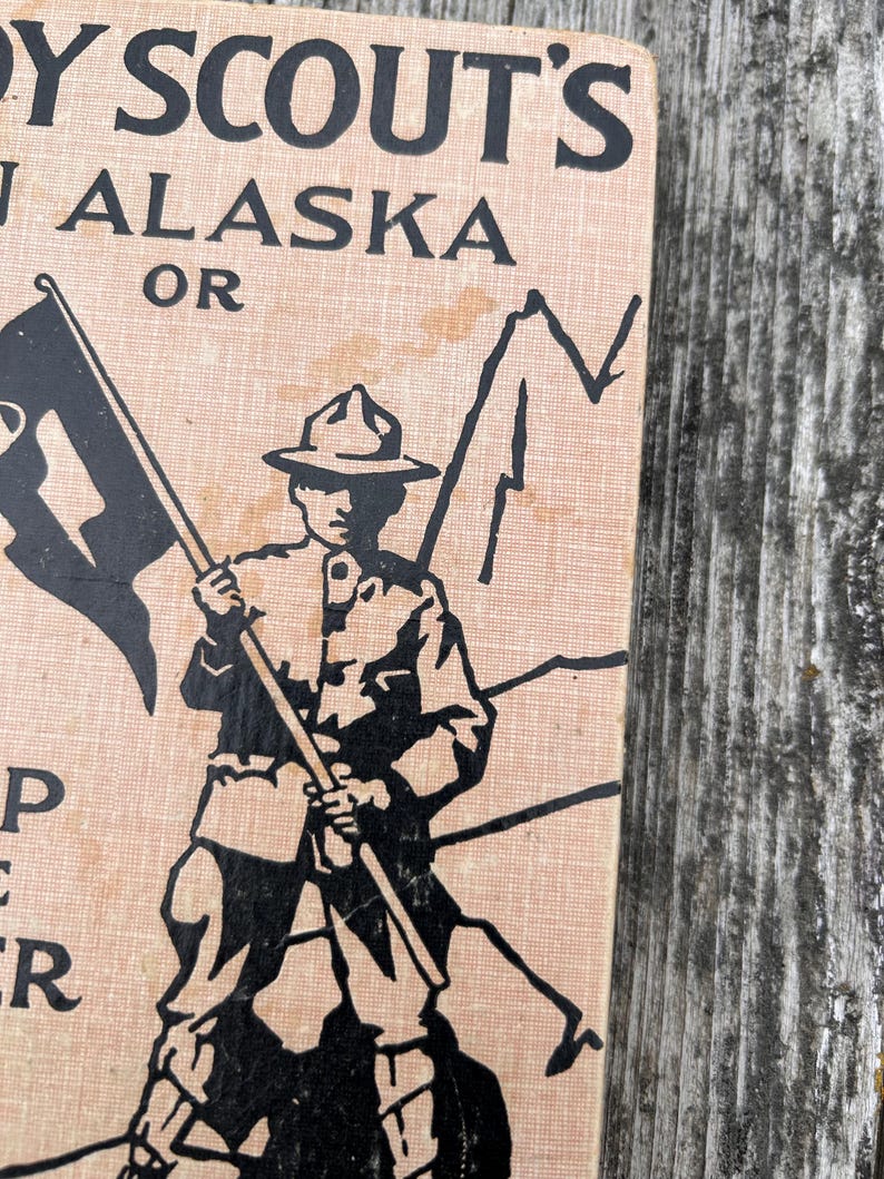 K&ouml;nnte beinhalten: Ein Vintage-Buchcover mit einem hellbraunen, strukturierten Hintergrund und schwarzen Tintenillustrationen. Der Text lautet BOY SCOUT'S IN ALASKA OR. Eine zentrale Grafik zeigt einen Pfadfinder mit breitkrempigem Hut, der einen Fahnenmast h&auml;lt.