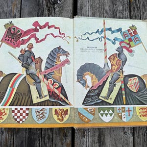 Może przedstawiać: Otwarta książka z kolorową ilustracją dw&oacute;ch rycerzy na koniach, z kt&oacute;rych każdy trzyma flagę i tarczę. Tekst "PROPERTY OF WALLOWA PUBLIC SCHOOLS DISTRICT #12 - WALLOWA, ORE." jest wydrukowany na prawej stronie nad postacią rycerza.