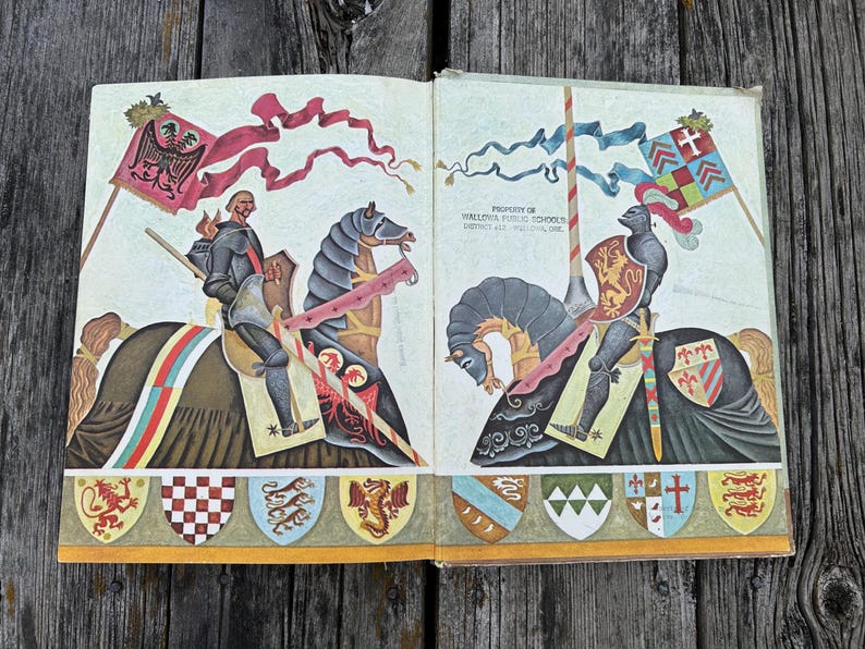 Może przedstawiać: Otwarta książka z kolorową ilustracją dw&oacute;ch rycerzy na koniach, z kt&oacute;rych każdy trzyma flagę i tarczę. Tekst "PROPERTY OF WALLOWA PUBLIC SCHOOLS DISTRICT #12 - WALLOWA, ORE." jest wydrukowany na prawej stronie nad postacią rycerza.