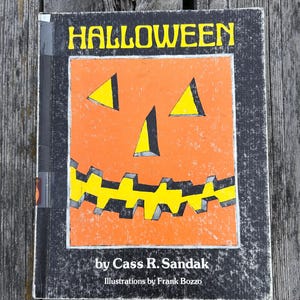 May include: A hardback book titled HALLOWEEN by Cass R. Sandak with illustrations by Frank Bozzo. The cover features a large orange jack-o'-lantern face with yellow triangular eyes and a jagged yellow mouth set against a black background.