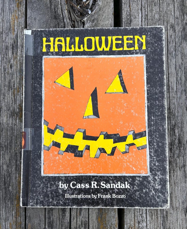 May include: A hardback book titled HALLOWEEN by Cass R. Sandak with illustrations by Frank Bozzo. The cover features a large orange jack-o'-lantern face with yellow triangular eyes and a jagged yellow mouth set against a black background.