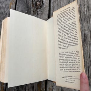 K&ouml;nnte beinhalten: Ein gebundenes Buch mit aufgeklappter Schutzumschlagklappe, die gedruckten biografischen Text &uuml;ber den Autor Alex Haley zeigt. Die cremefarbenen Papierseiten sind sichtbar und der Text enth&auml;lt Details &uuml;ber das Buch Roots und die Karriere des Autors.