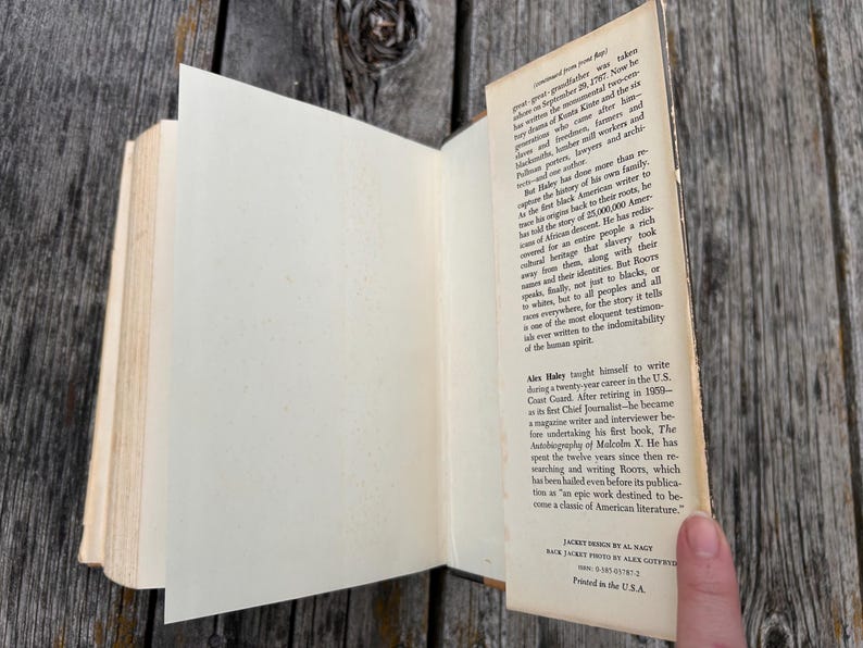 K&ouml;nnte beinhalten: Ein gebundenes Buch mit aufgeklappter Schutzumschlagklappe, die gedruckten biografischen Text &uuml;ber den Autor Alex Haley zeigt. Die cremefarbenen Papierseiten sind sichtbar und der Text enth&auml;lt Details &uuml;ber das Buch Roots und die Karriere des Autors.