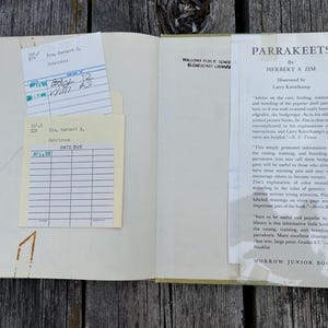May include: An open hardback book titled Parrakeets by Herbert S. Zim, featuring a library checkout pocket with two cream-coloured date due cards. The right page displays a printed book jacket flap with text about parrakeet care and breeding.