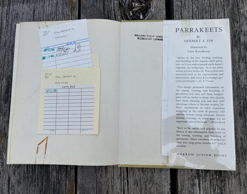 May include: An open hardback book titled Parrakeets by Herbert S. Zim, featuring a library checkout pocket with two cream-coloured date due cards. The right page displays a printed book jacket flap with text about parrakeet care and breeding.