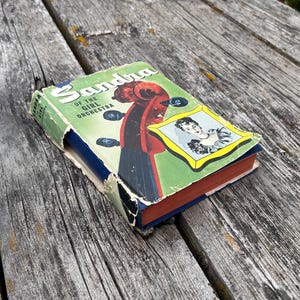May include: A vintage hardback book titled Sandra of the Girl Orchestra with a green dust jacket featuring a red violin scroll, black tuning pegs, and a framed illustration of a person. The book has worn edges and a blue spine with red page edges.
