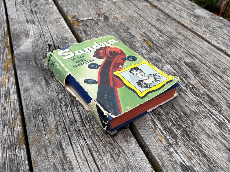 May include: A vintage hardback book titled Sandra of the Girl Orchestra with a green dust jacket featuring a red violin scroll, black tuning pegs, and a framed illustration of a person. The book has worn edges and a blue spine with red page edges.