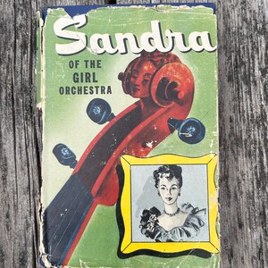 May include: A vintage book titled Sandra of the Girl Orchestra with a green cover featuring a red cello scroll and a yellow-framed illustration of a person wearing a ruffled top and a necklace. The book shows signs of wear along the edges.