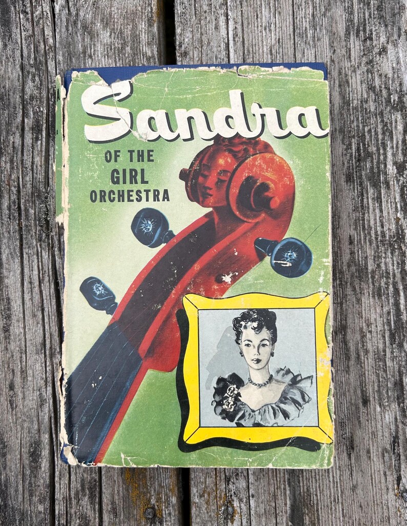 May include: A vintage book titled Sandra of the Girl Orchestra with a green cover featuring a red cello scroll and a yellow-framed illustration of a person wearing a ruffled top and a necklace. The book shows signs of wear along the edges.