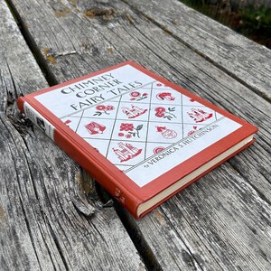 May include: A hardcover book titled Chimney Corner Fairy Tales by Veronica S. Hutchinson with a burnt orange spine and edges. The front cover features a white diamond grid pattern with red illustrations of flowers, castles, and teapots.