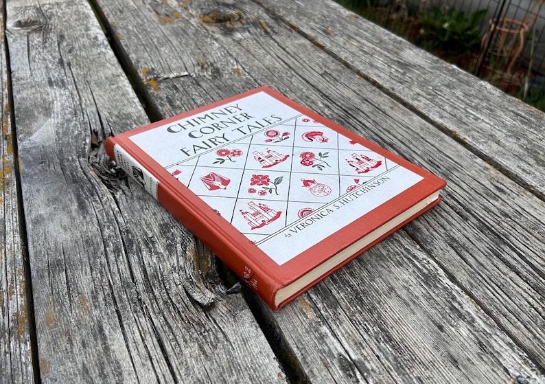 May include: A hardcover book titled Chimney Corner Fairy Tales by Veronica S. Hutchinson with a burnt orange spine and edges. The front cover features a white diamond grid pattern with red illustrations of flowers, castles, and teapots.