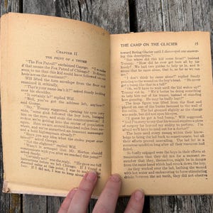K&ouml;nnte beinhalten: Ein aufgeschlagenes altes Buch mit vergilbten Seiten, das links Kapitel II mit dem Titel The Print of a Thumb und rechts The Camp on the Glacier zeigt. Der Text ist in schwarzer Tinte auf gealtertem Papier mit sichtbarem Verschlei&szlig; gedruckt.