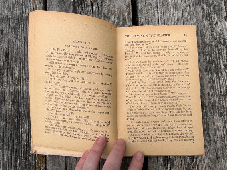 K&ouml;nnte beinhalten: Ein aufgeschlagenes altes Buch mit vergilbten Seiten, das links Kapitel II mit dem Titel The Print of a Thumb und rechts The Camp on the Glacier zeigt. Der Text ist in schwarzer Tinte auf gealtertem Papier mit sichtbarem Verschlei&szlig; gedruckt.