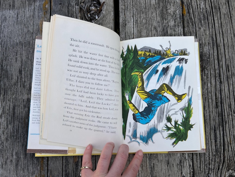 Pu&ograve; includere: Un libro con copertina rigida aperto che presenta testo stampato in nero sulla pagina sinistra e un'illustrazione colorata sulla destra. L'immagine mostra una figura con una tunica blu e leggings gialli che cade da una cascata tra gli alberi.