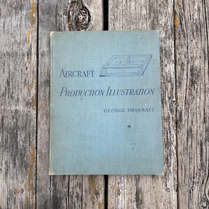 Pu&ograve; includere: Un libro con copertina rigida intitolato Aircraft Production Illustration di George Tharratt con una copertina azzurra. La copertina presenta un disegno al tratto blu scuro di un tavolo da disegno con una riga a T e una squadra, oltre al titolo e l'autore.
