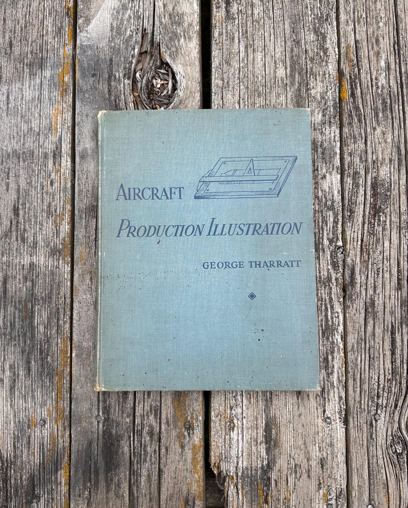 Pu&ograve; includere: Un libro con copertina rigida intitolato Aircraft Production Illustration di George Tharratt con una copertina azzurra. La copertina presenta un disegno al tratto blu scuro di un tavolo da disegno con una riga a T e una squadra, oltre al titolo e l'autore.