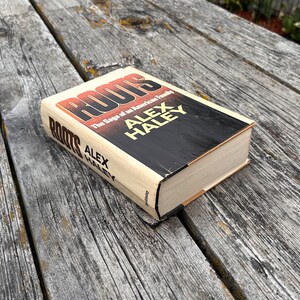 K&ouml;nnte beinhalten: Ein gebundenes Buch mit dem Titel Roots: The Saga of an American Family von Alex Haley. Der Einband zeigt eine markante rote und schwarze Schrift auf cremefarbenem Hintergrund, wobei der Name des Autors in gro&szlig;en gelben Buchstaben auf der Vorderseite und dem Buchr&uuml;cken steht.
