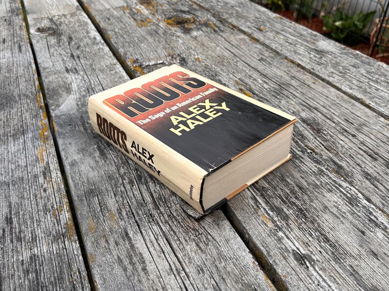 K&ouml;nnte beinhalten: Ein gebundenes Buch mit dem Titel Roots: The Saga of an American Family von Alex Haley. Der Einband zeigt eine markante rote und schwarze Schrift auf cremefarbenem Hintergrund, wobei der Name des Autors in gro&szlig;en gelben Buchstaben auf der Vorderseite und dem Buchr&uuml;cken steht.
