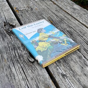 Pu&ograve; includere: Un libro con copertina rigida intitolato Leif the Lucky di Erick Berry, un Discovery Book. La copertina presenta uno sfondo blu e bianco con l'illustrazione di una persona in tunica gialla e pantaloni verdi, con spada ed elmo visibili.