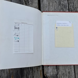 May include: An open hardcover book with a red spine and white endpapers. The left side features a white library date due card with various stamped dates. The right side contains a pale yellow library checkout card with typed text and a call number.