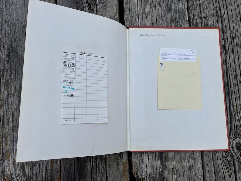 May include: An open hardcover book with a red spine and white endpapers. The left side features a white library date due card with various stamped dates. The right side contains a pale yellow library checkout card with typed text and a call number.
