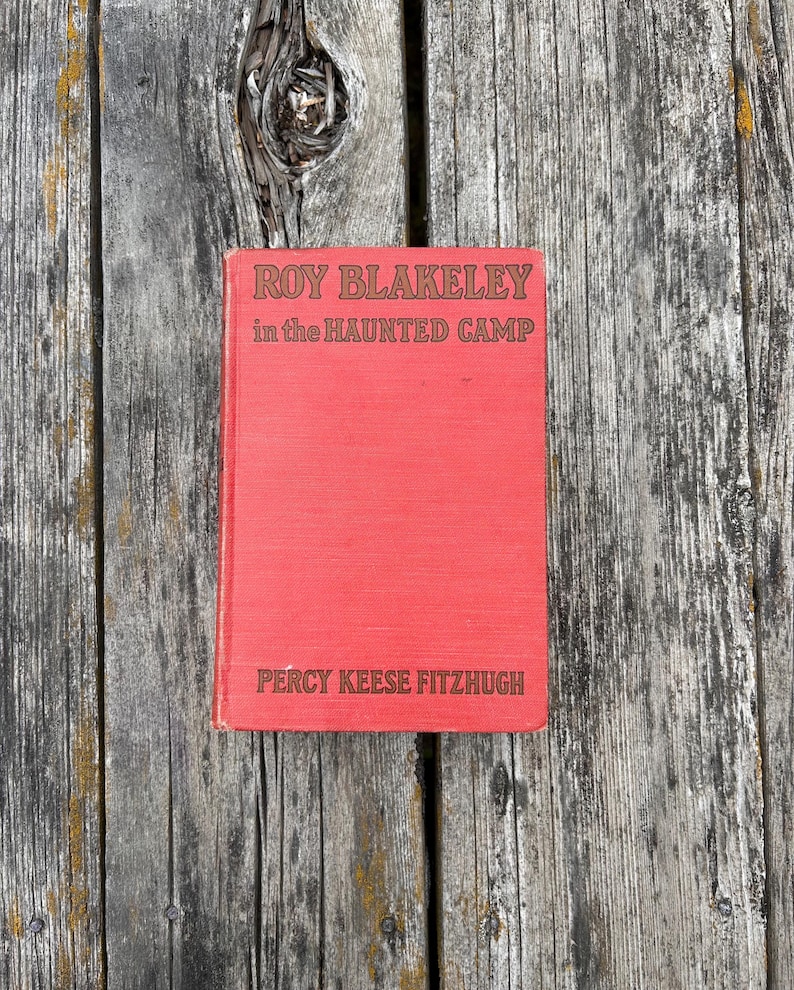 May include: A red hardcover book titled Roy Blakeley in the Haunted Camp by Percy Keese Fitzhugh. The front cover features dark brown embossed lettering against a textured red fabric background, displaying the title and author name clearly.