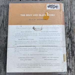 May include: The back cover of a hardcover book titled The Billy and Blaze Books by C. W. Anderson. The cover is tan and white with black text listing book titles and summaries. It features a library sticker at the top and a torn yellow label.