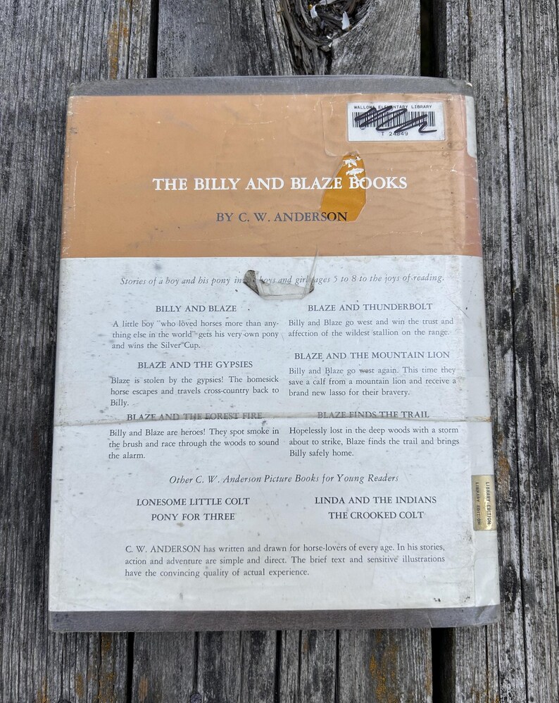 May include: The back cover of a hardcover book titled The Billy and Blaze Books by C. W. Anderson. The cover is tan and white with black text listing book titles and summaries. It features a library sticker at the top and a torn yellow label.