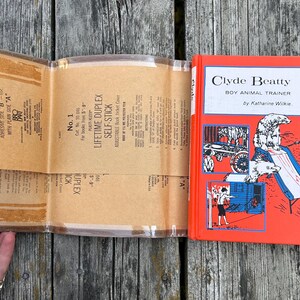 May include: A hardback book titled Clyde Beatty Boy Animal Trainer by Katherine Wilkie with a bright red cover featuring illustrations of a trainer and polar bears, accompanied by a clear, adhesive plastic book jacket cover with printed instructions.