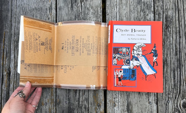 May include: A hardback book titled Clyde Beatty Boy Animal Trainer by Katherine Wilkie with a bright red cover featuring illustrations of a trainer and polar bears, accompanied by a clear, adhesive plastic book jacket cover with printed instructions.