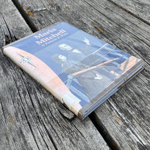Puede incluir: Un libro biogr&aacute;fico de tapa dura titulado Maria Mitchell por Katharine E. Wilkie, con una cubierta azul que presenta una ilustraci&oacute;n blanca de una persona mirando a trav&eacute;s de un telescopio y una rosa de los vientos a la izquierda, en una funda pl&aacute;stica.