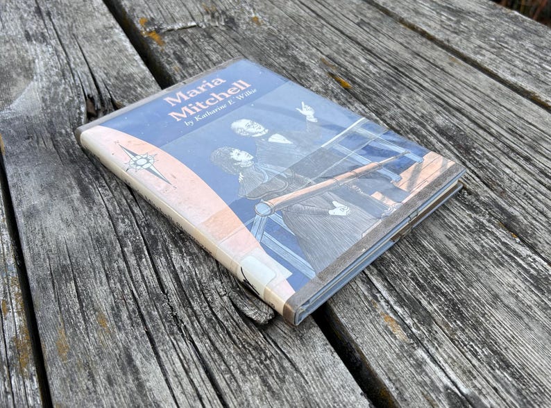 Puede incluir: Un libro biogr&aacute;fico de tapa dura titulado Maria Mitchell por Katharine E. Wilkie, con una cubierta azul que presenta una ilustraci&oacute;n blanca de una persona mirando a trav&eacute;s de un telescopio y una rosa de los vientos a la izquierda, en una funda pl&aacute;stica.