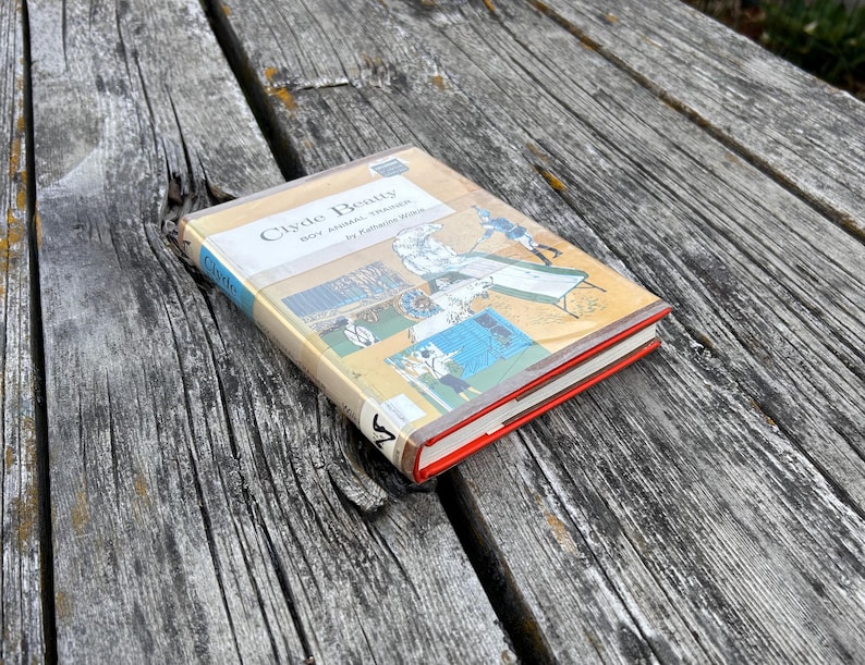 May include: A hardback book titled Clyde Beatty Boy Animal Trainer by Katherine Wilkie. The cover features a tan background with a blue spine and a graphic illustration of a person training animals. The book has a bright red edge on the pages.