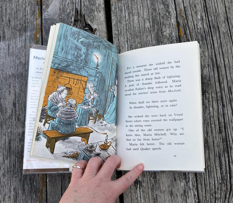 Puede incluir: Un libro de tapa dura abierto que presenta una ilustraci&oacute;n en color de tres mujeres sentadas junto a una chimenea en la p&aacute;gina izquierda y texto impreso en la derecha. El texto incluye las l&iacute;neas: When shall we three meet again, In thunder, lightning, or in rain?