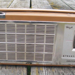 Może przedstawiać: Prostokątne radio Oritone 8 Transistor z obudową w kolorze brązowym przypominającym sk&oacute;rę oraz srebrnym metalowym panelem przednim. Urządzenie posiada poziomą kratkę głośnika, g&oacute;rny uchwyt do przenoszenia oraz boczne pokrętło z napisem High Fidelity.