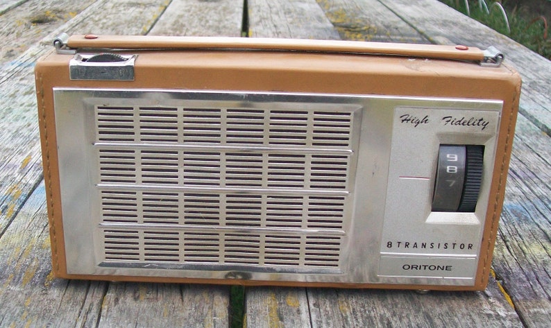 Może przedstawiać: Prostokątne radio Oritone 8 Transistor z obudową w kolorze brązowym przypominającym sk&oacute;rę oraz srebrnym metalowym panelem przednim. Urządzenie posiada poziomą kratkę głośnika, g&oacute;rny uchwyt do przenoszenia oraz boczne pokrętło z napisem High Fidelity.