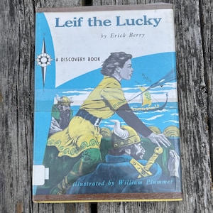 Pu&ograve; includere: Un libro con copertina rigida intitolato Leif the Lucky di Erick Berry, illustrato da William Plummer. La copertina presenta un'illustrazione blu, gialla e verde di figure in abiti vichinghi su una nave, con una rosa dei venti e il testo A Discovery Book.