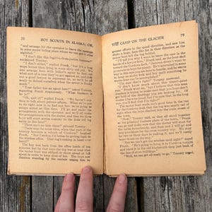 K&ouml;nnte beinhalten: Ein aufgeschlagenes altes Buch mit vergilbten Seiten, das die Seiten 78 und 79 zeigt. Die linke Seite ist mit Boy Scouts in Alaska; or, und die rechte mit The Camp on the Glacier betitelt. Der Text ist in schwarzer Tinte mit Abs&auml;tzen gedruckt.