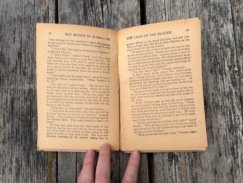 K&ouml;nnte beinhalten: Ein aufgeschlagenes altes Buch mit vergilbten Seiten, das die Seiten 78 und 79 zeigt. Die linke Seite ist mit Boy Scouts in Alaska; or, und die rechte mit The Camp on the Glacier betitelt. Der Text ist in schwarzer Tinte mit Abs&auml;tzen gedruckt.
