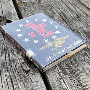 May include: A hardcover book titled The Story of Our Flag by Carl Glick and Ollie Rogers. The dark blue cover features a circular arrangement of white stars, a gold eagle emblem, and red text. The book spine also displays the title.