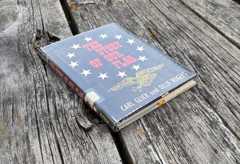 May include: A hardcover book titled The Story of Our Flag by Carl Glick and Ollie Rogers. The dark blue cover features a circular arrangement of white stars, a gold eagle emblem, and red text. The book spine also displays the title.