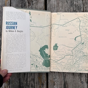 May include: A hardback book titled Russian Journey by William O. Douglas, featuring a cream-coloured dust jacket with blue text and a green map illustration of the Soviet Union on the inside cover, displaying geographical details and borders.