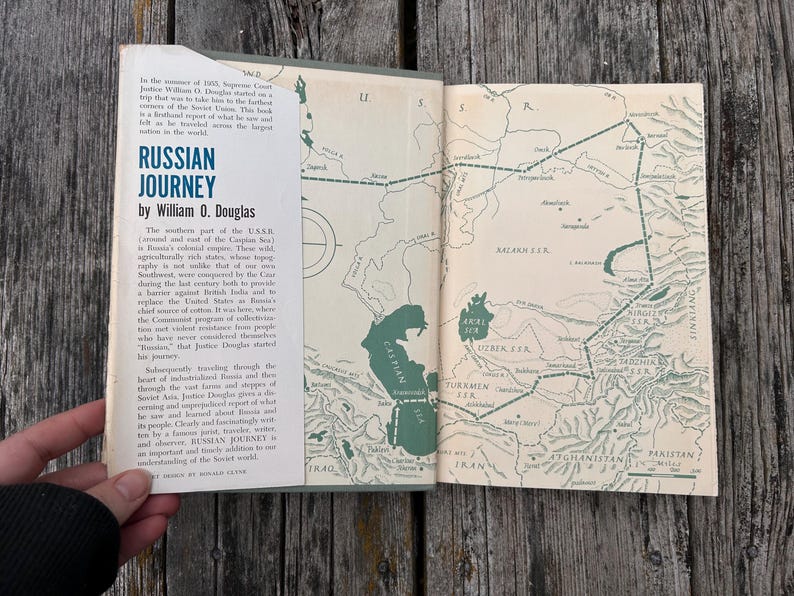 May include: A hardback book titled Russian Journey by William O. Douglas, featuring a cream-coloured dust jacket with blue text and a green map illustration of the Soviet Union on the inside cover, displaying geographical details and borders.