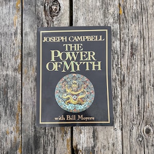 K&ouml;nnte beinhalten: Ein gebundenes Buch mit dem Titel The Power of Myth von Joseph Campbell mit Bill Moyers. Der schwarze Einband zeigt goldenen Text und eine kreisf&ouml;rmige, farbenfrohe Drachenillustration unter dem Titel, umrahmt von einer d&uuml;nnen Goldlinie.
