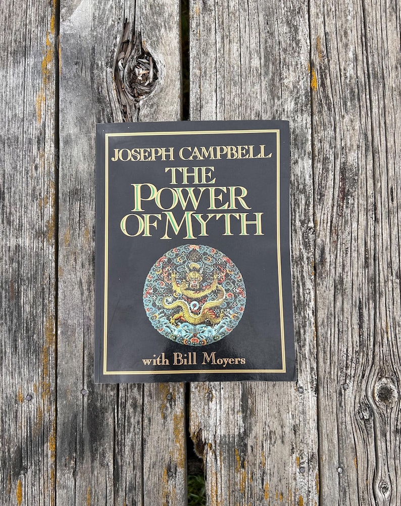 K&ouml;nnte beinhalten: Ein gebundenes Buch mit dem Titel The Power of Myth von Joseph Campbell mit Bill Moyers. Der schwarze Einband zeigt goldenen Text und eine kreisf&ouml;rmige, farbenfrohe Drachenillustration unter dem Titel, umrahmt von einer d&uuml;nnen Goldlinie.