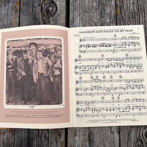 K&ouml;nnte beinhalten: Ein aufgeschlagenes Musikbuch mit den Noten f&uuml;r Raindrops Keep Fallin' On My Head. Die linke Seite zeigt ein Schwarz-Wei&szlig;-Foto von f&uuml;nf M&auml;nnern in Westernkleidung, w&auml;hrend die rechte Seite die musikalische Notation und den Liedtext enth&auml;lt.