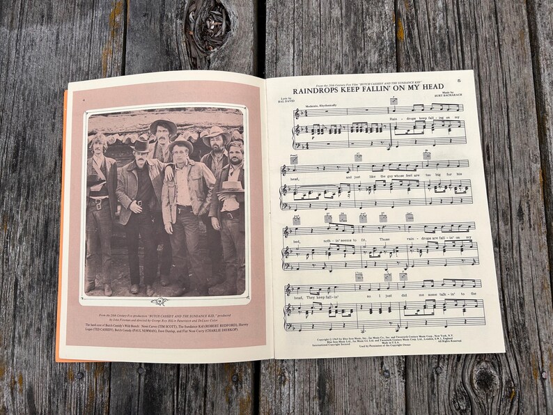 K&ouml;nnte beinhalten: Ein aufgeschlagenes Musikbuch mit den Noten f&uuml;r Raindrops Keep Fallin' On My Head. Die linke Seite zeigt ein Schwarz-Wei&szlig;-Foto von f&uuml;nf M&auml;nnern in Westernkleidung, w&auml;hrend die rechte Seite die musikalische Notation und den Liedtext enth&auml;lt.