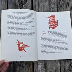 Może przedstawiać: Otwarta książka z czarnym tekstem na białych stronach, zawierająca dwie ilustracje czerwono-białych tarcz herbowych. Lewa strona jest oznaczona numerem 14, a prawa numerem 15, z widocznym tytułem The Knighting of Arthur.